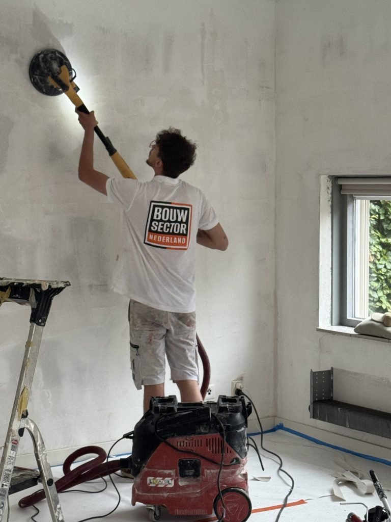 Vakman van Bouwsector Nederland schuurt een wand met een schuurmachine op een steel, de vloer is afgedekt