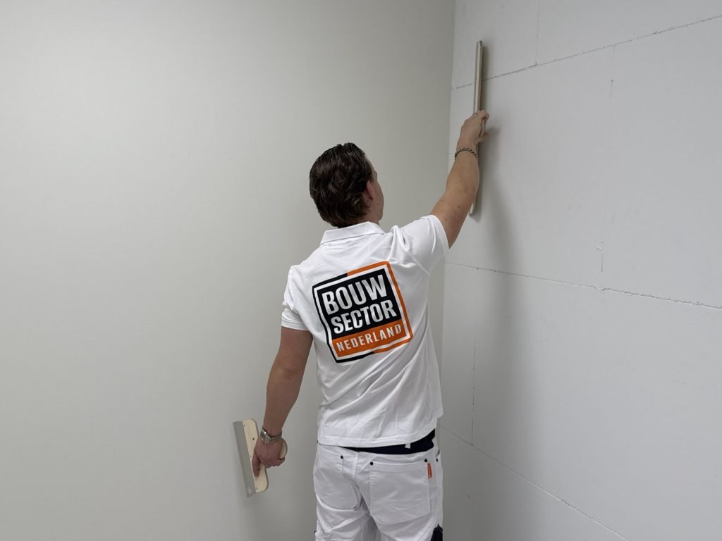 Stukadoor van Bouwsector Nederland houdt een lange spaan verticaal tegen een wand, met een tweede spaan in zijn andere hand