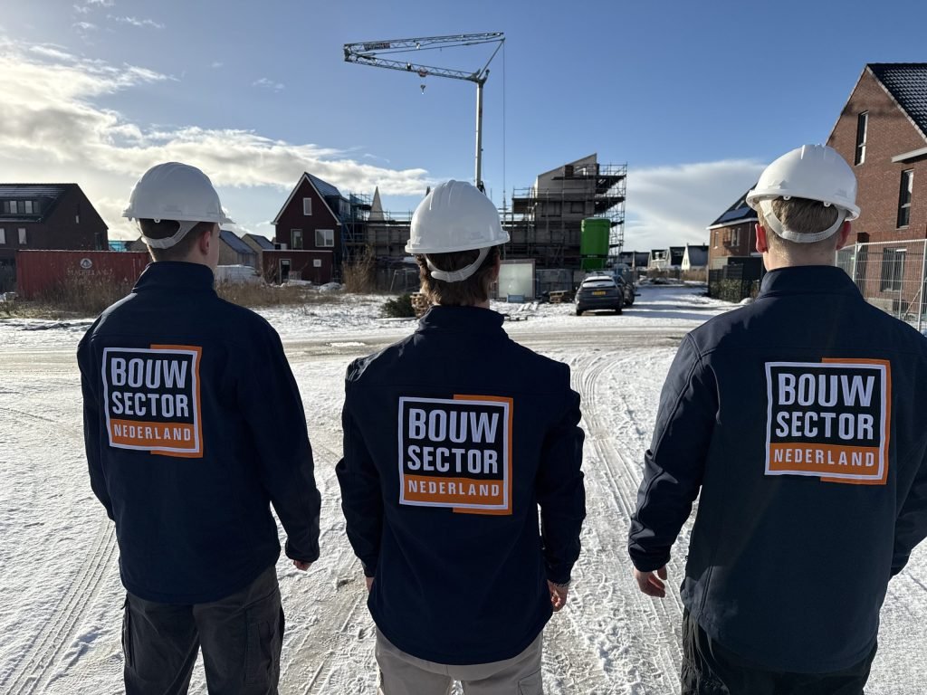 Team in donkerblauwe jassen met oranje logo op de rug voor een bouwplaats onder een blauwe lucht
